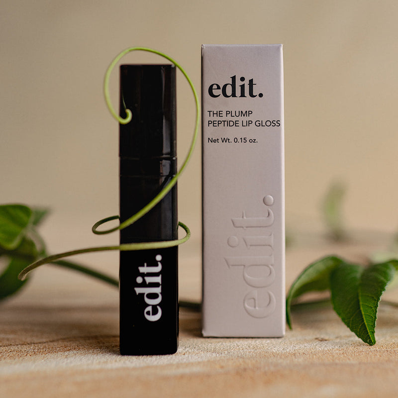 Black lip gloss with 'edit.' branding next to its packaging on a wooden surface with leaves in the background.