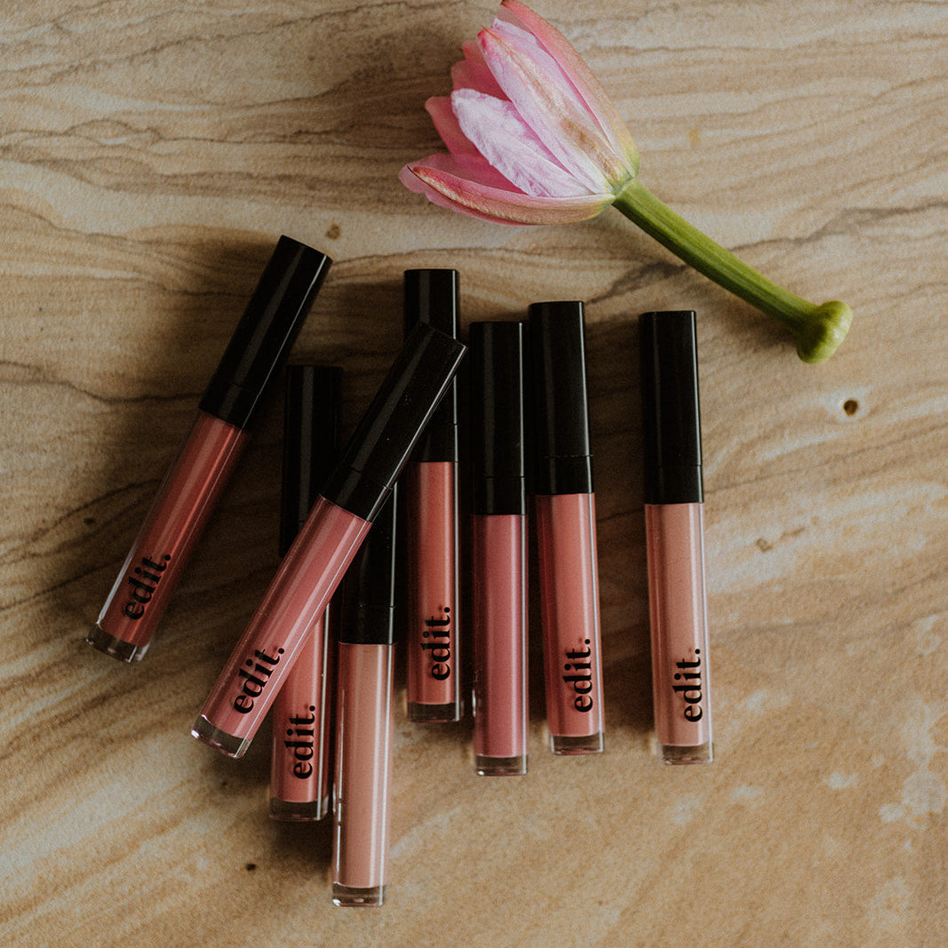 Set of lip glosses with black caps on a wooden surface next to a pink flower.