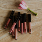 Set of lip glosses with black caps on a wooden surface next to a pink flower.