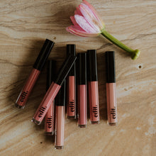 Set of lip glosses with black caps on a wooden surface next to a pink flower.